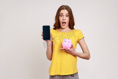 身穿黄色休闲风格 T 恤、棕色头发、手持存钱罐和带空白屏幕广告的智能手机的惊讶女性肖像。