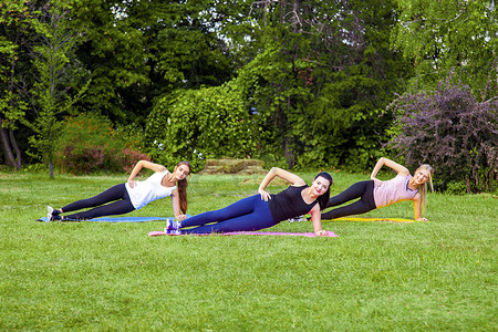 三个美丽健康、黏糊糊的女人在公园的绿草地上做运动，侧着双臂，带着露齿的微笑看着镜头。