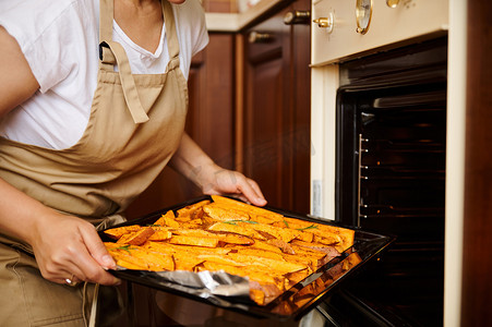 一位穿着厨师围裙的家庭主妇的特写镜头，将一张带有生有机红薯块的烤盘放入烤箱中