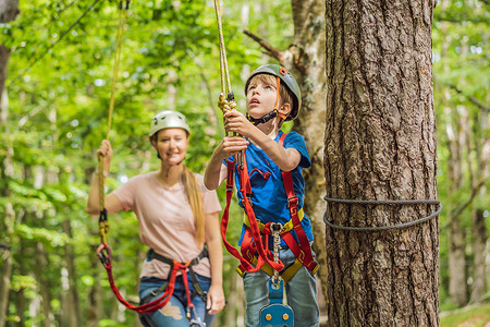 树顶摄影照片_母亲和儿子在森林里的极限公路电车滑索上攀登树上的登山绳安全连接到树顶绳索冒险公园。