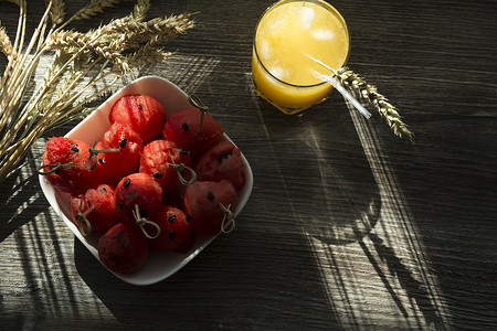 红色果汁摄影照片_阳光下的木桌上放着鲜榨橙汁、甜西瓜甜点和成熟小麦的小穗。