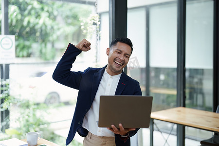 幸福感摄影照片_一位亚洲男性企业主站在电脑前的肖像，在成功投资后表现出幸福感