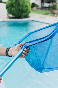 手握着背景为蓝色水池的撇渣器，一名男子用撇叶器清洁泳池。