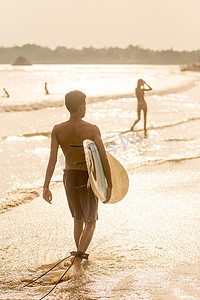 阳光下散步摄影照片_阳光下，一名男子手持冲浪板在斯里兰卡米迪加马热带海滩散步。