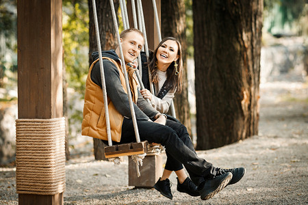 秋天，一对可爱的年轻夫妇在公园户外穿着基本颜色的温暖衣服骑着秋千的侧视图