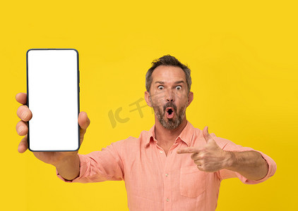 巨大手机摄影照片_惊讶的中年灰发男子用手指指着智能手机，看着穿着黄色隔离桃色衬衫的相机。