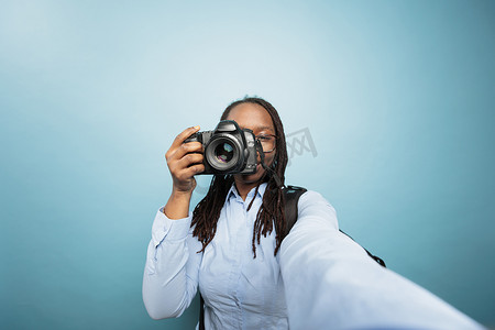 单反图片摄影照片_年轻的非洲裔美国成年女性在蓝色背景下拍照时将数码单反设备指向相机。