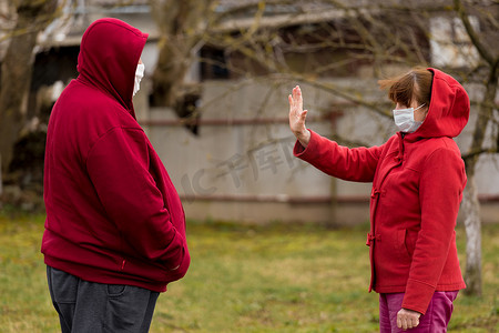 一位戴着医用防护口罩的老妇人站在户外街道前，做出了与老人保持社交距离的手势
