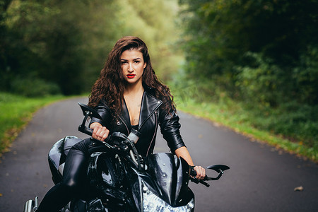 冷淡摄影照片_一位美丽的年轻女子骑着摩托车，穿着黑色皮衣，在大自然、街道、森林里，在阳光明媚的天气里，特写，看着相机的肖像
