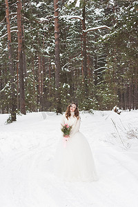 美丽的新娘穿着白色连衣裙，在冰雪覆盖的冬季森林里捧着花束。