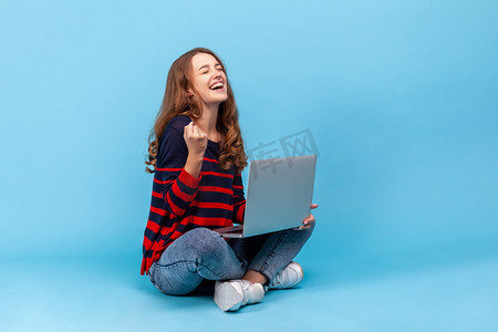 女人坐在膝盖上，笔记本电脑放在膝盖上，表达幸福，庆祝她的成功，握紧拳头。