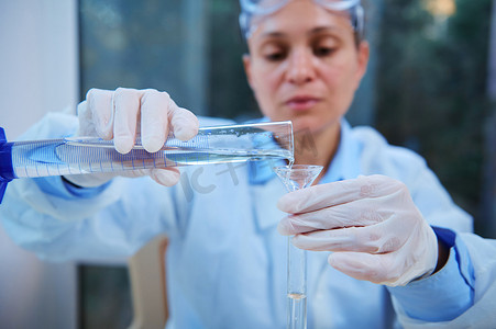 制药公司摄影照片_细节：一位模糊科学家手中的量筒，通过实验室漏斗将液体倒入试管