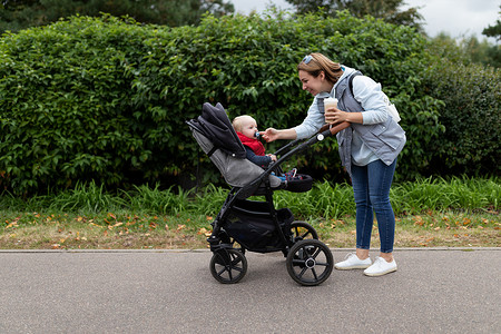 妈妈推孩子摄影照片_一位年轻的母亲手里拿着一杯咖啡，脸上带着微笑，在公园里推着婴儿车走向她的小宝宝时，伸直了乳头