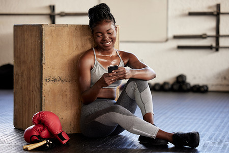 运动妇女摄影照片_电话、健身和应用程序，一位黑人女拳击手在健身房通过移动应用程序跟踪她的锻炼、锻炼和进度。