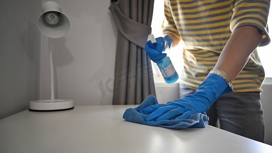 戴着橡胶手套的男子用抹布和喷雾清洁剂擦拭白桌上灰尘的裁剪图像。