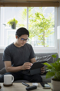 家庭用电摄影照片_年轻的亚洲男子坐在沙发上，用电脑平板电脑在线工作。