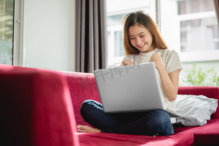 年轻女子在红色沙发上用笔记本电脑上网，很高兴在她的房子里成功进行交易。