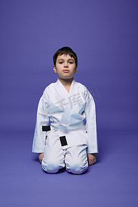 儿童跆拳道摄影照片_一个穿着和服、坐在紫色背景下的合气道姿势、带有文本复制空间的英俊男孩的肖像。