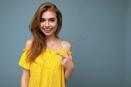 文字夏天摄影照片_照片中，年轻可爱、美丽、迷人、微笑、积极、快乐的深色金发女人，在蓝色背景墙上隔离着真诚的情感，并有复制空间，可供穿着时尚黄色连衣裙的文字。