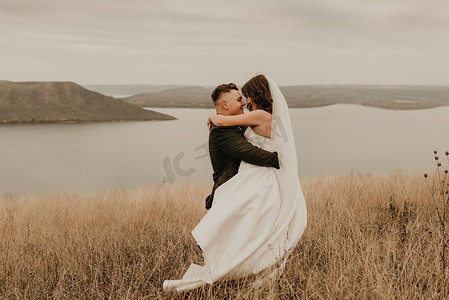 恩爱夫妻的婚礼，身穿礼服面纱、运动鞋和西装的新婚夫妇拥抱亲吻，在河上山田里的高高的草地上旋转