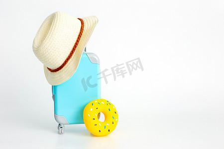 白色背景上站着一个带轮子的蓝色手提箱、一顶草帽和一个黄色充气甜甜圈。