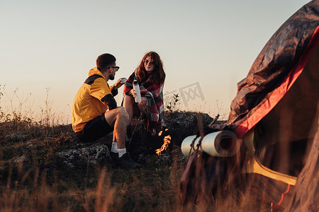 男女喝摄影照片_日落时，两个年轻的旅行者男女坐在户外的篝火和帐篷附近，喝着热水瓶里的热茶