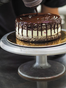 融化的巧克力滴摄影照片_厨师蛋糕装饰师将装有巧克力甘纳许的瓶子挤在蛋糕上