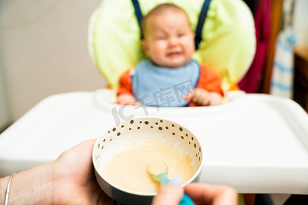 抗住饥饿摄影照片_坐在高脚椅上的宝宝不肯吃妈妈用勺子给的食物，调皮、哭闹。