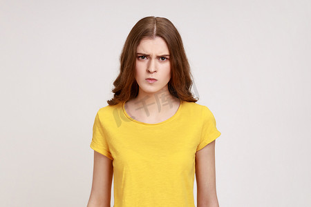 年轻美丽的非常愤怒的少女穿着黄色 T 恤，棕色波浪发，直视镜头，面部表情严格。