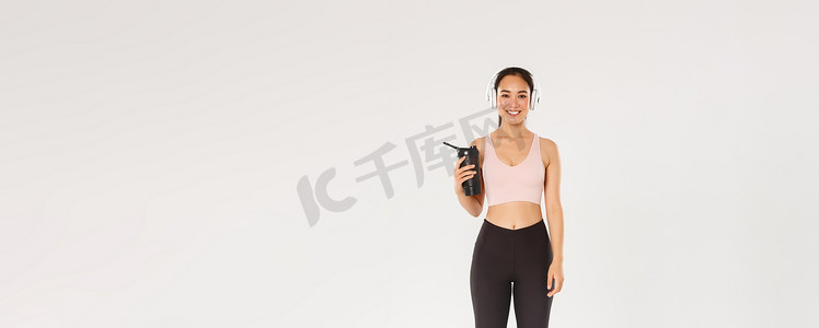 全长微笑的苗条而快乐的健身女孩，亚洲女运动员戴着耳机在健身房锻炼，锻炼期间听音乐，锻炼之间从瓶子里喝水，白色背景