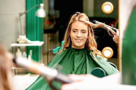 婚礼用机摄影照片_理发师用卷发钳在美容院为年轻的白人女孩卷曲长长的棕色头发。