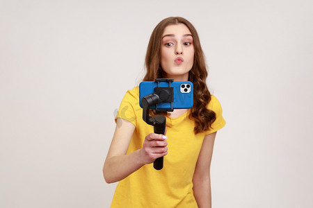 年轻漂亮的女人，带着稳定器上的拍照手机，为她的视频博客拍摄视频，做出接吻的手势，穿着黄色休闲风格的 T 恤。