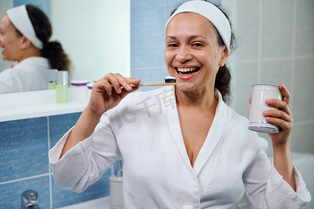 身穿白色浴袍的多民族妇女用生态竹牙刷和美白牙膏刷牙，对着镜头微笑