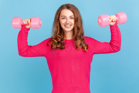 健身教练摄影照片_女人拿着粉色哑铃看着相机，在健身房里锻炼二头肌。