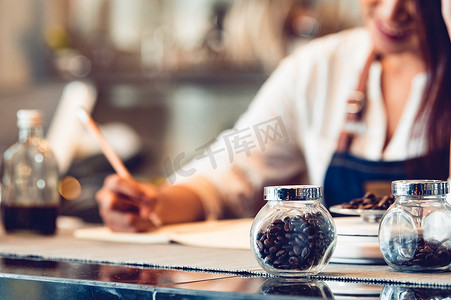 咖啡豆瓶和快乐女咖啡师的特写在笔记本上写下报告笔记，以计算作为咖啡店店主企业家背景的业务成本和利润。