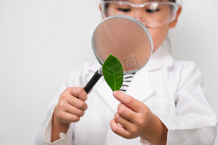 一个戴着眼镜的小女孩拿着放大镜看着白色背景上穿着研究员或科学制服的叶子的肖像。