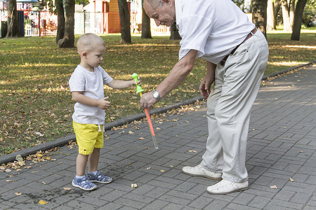 带孩子的老人摄影照片_周末，一位老爷爷带着孙子在游乐园里玩耍，兴高采烈地吹着肥皂泡。