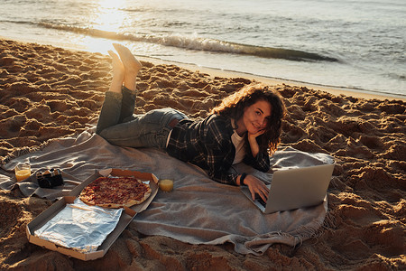 黎明时分躺在海边，快乐的卷发女人一边吃披萨，一边在笔记本电脑上工作