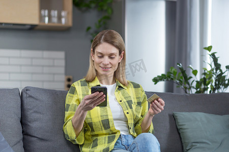 快乐的女人坐在家里的沙发上，使用电话和银行信用卡在网上购物