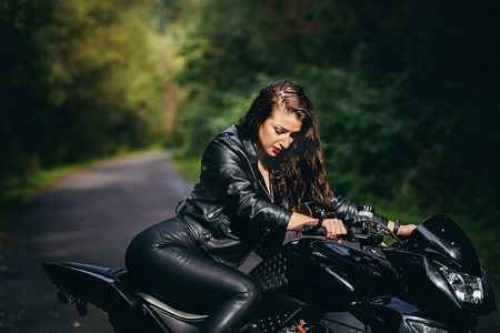 一位美丽的年轻女子骑着摩托车，穿着黑色皮衣，在大自然、街道、森林里，在阳光明媚的天气里，特写，看着相机的肖像