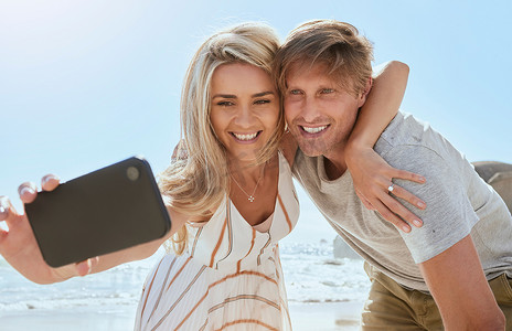 恩爱的年轻夫妇一边用手机自拍，一边微笑和拥抱在海滩享受一天。