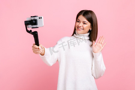 女博主摄影照片_快乐的女博主使用智能手机和斯坦尼康来进行流媒体、挥手、问候追随者。