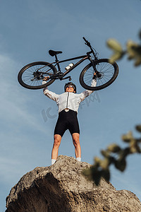 职业男自行车手站在山顶，背景是天空，举起自行车双手