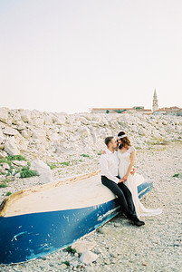 海滩婚礼摄影照片_新娘站在新郎旁边，坐在岸边一艘倒置的渔船上