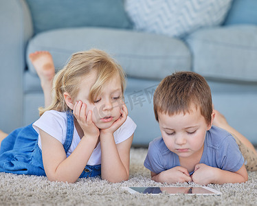 女孩卡通摄影照片_年轻的白人男孩和女孩躺在家里的地毯上，在线观看卡通视频，在数字平板电脑上玩有趣的游戏，从教育应用程序中学习。