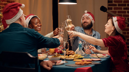 碰杯香槟摄影照片_圣诞晚餐上，戴着传统帽子、碰杯香槟的喜庆多元化人们