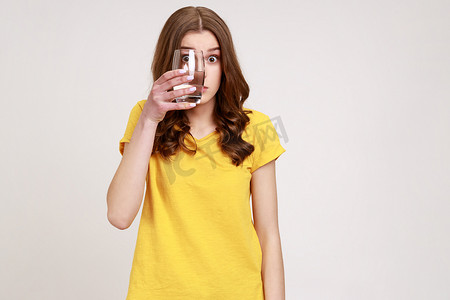 年轻漂亮的女性穿着黄色休闲 T 恤，用一杯水遮住眼睛，用惊讶的大眼睛看着投掷玻璃。