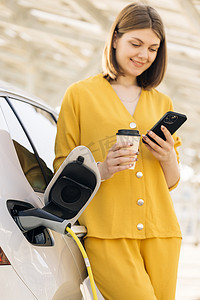 拿着手机和咖啡杯的白人妇女站在充电电动车附近。
