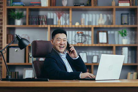 行政文员摄影照片_快乐的亚洲银行家老板在经典办公室里用笔记本电脑打电话