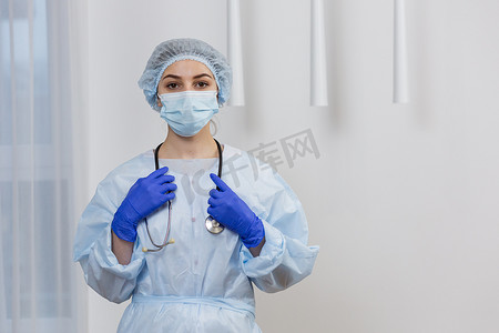 戴着面具、手术服和帽子的年轻女医生的肖像，站着听诊器，看着相机